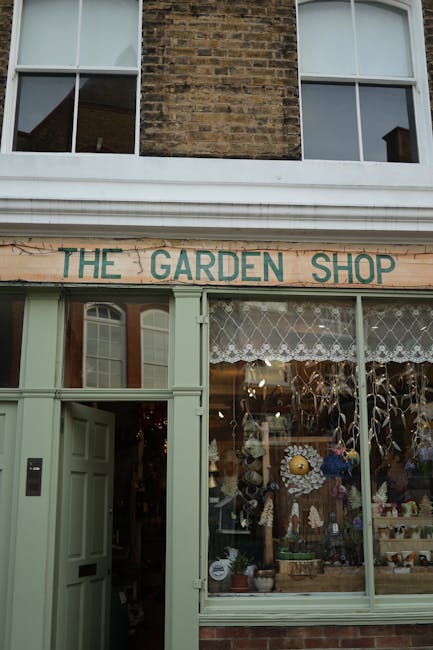 The exterior of a shop called 'The Garden Shop' features large glass windows revealing a variety of garden-related items and decorations inside, including potted plants, gardening ornaments, and flora