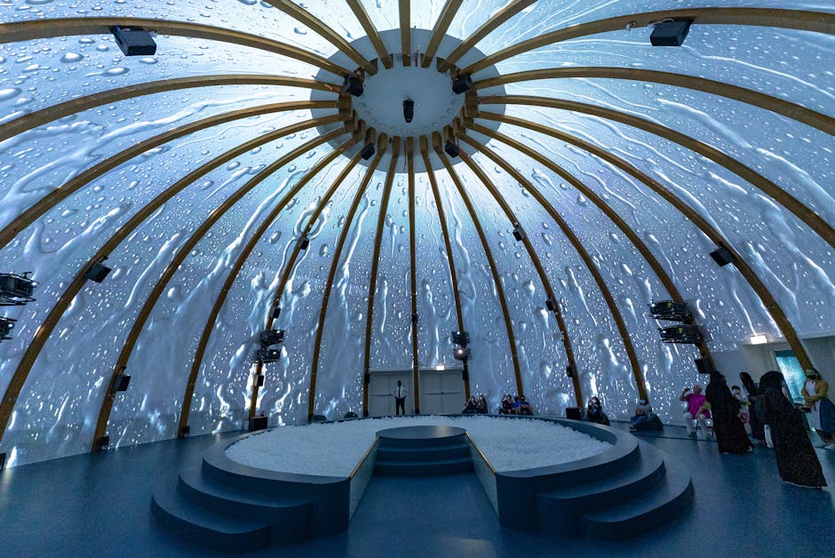 Inside a large, dome-shaped structure resembling a greenhouse or eco-education space, featuring a wooden framework with curved beams supporting a semi-transparent ceiling. The ceiling's surface is ado